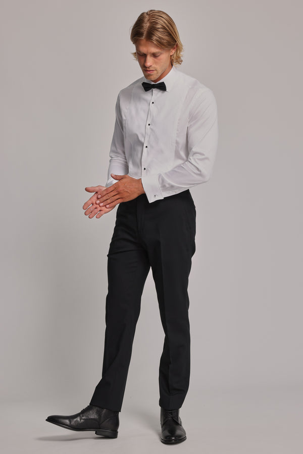 A man with blonde hair models the Barkers Eldridge Dress Shirt, featuring a tuxedo bib, paired with black dress pants, bow tie, and shoes. He stands against a plain background, looking down while adjusting his hands in elegant formal wear.