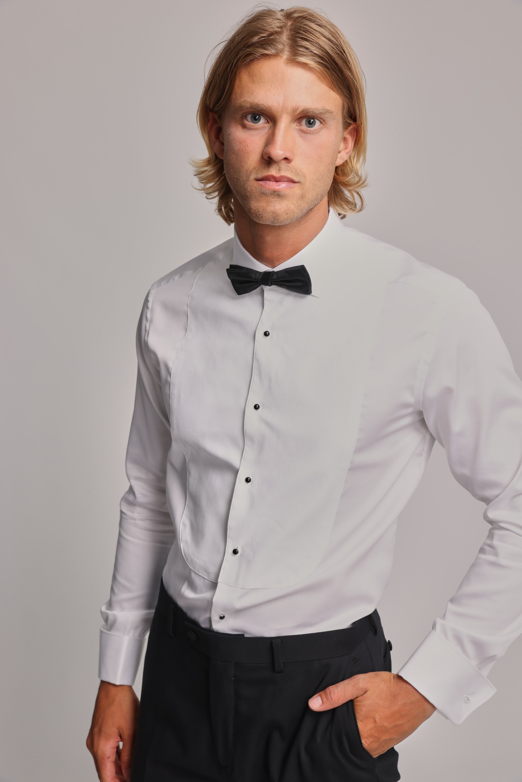 A young man with blond hair wears the Barkers Eldridge Dress Shirt, featuring a tuxedo bib, paired with a black bow tie and pants. He stands with one hand in his pocket, looking at the camera against a plain light background.