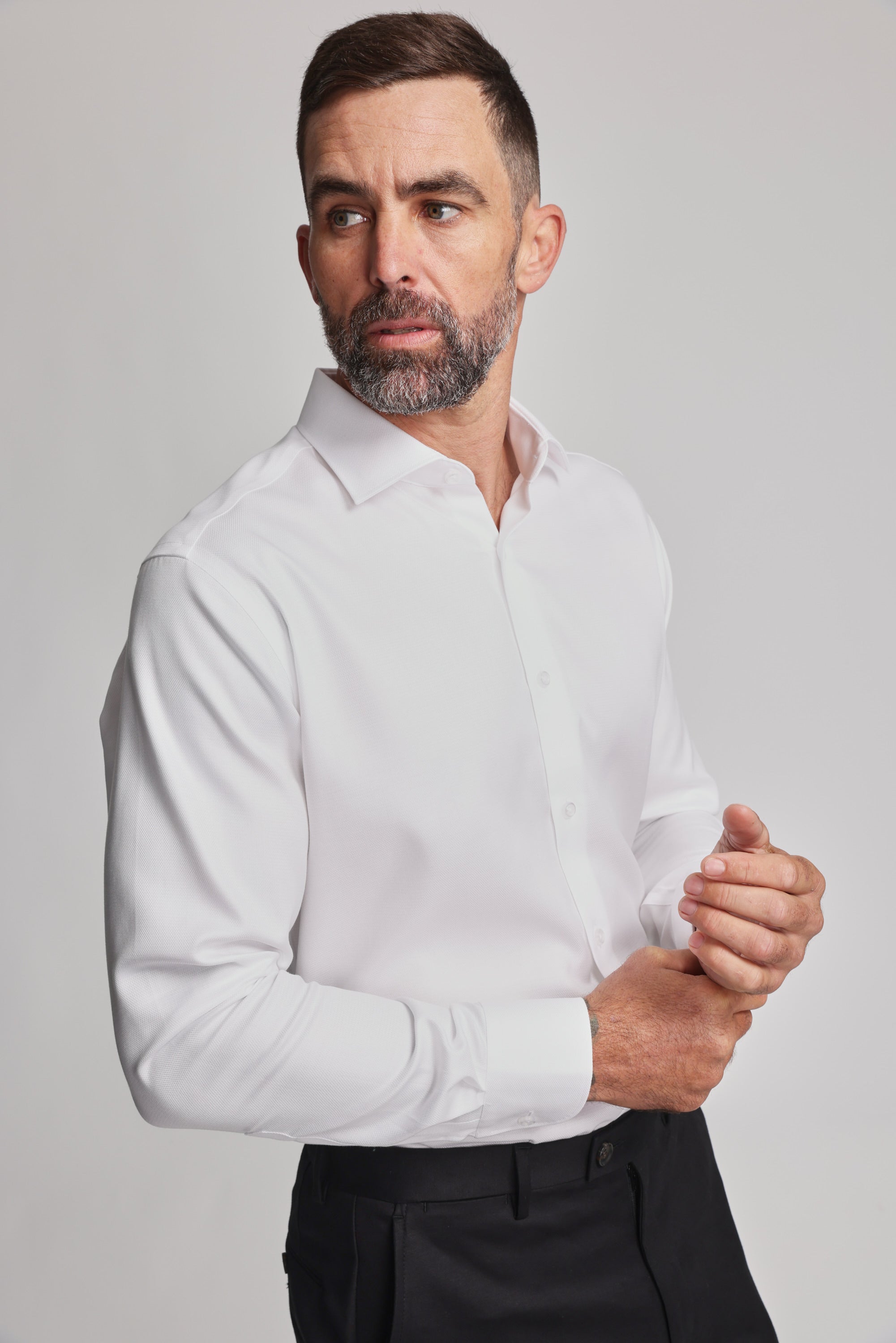 A man with short dark hair and a beard wears the Barkers Mexted Texture Shirt and black pants, looking to his left with a neutral expression while adjusting his shirt cuff against a plain light background.