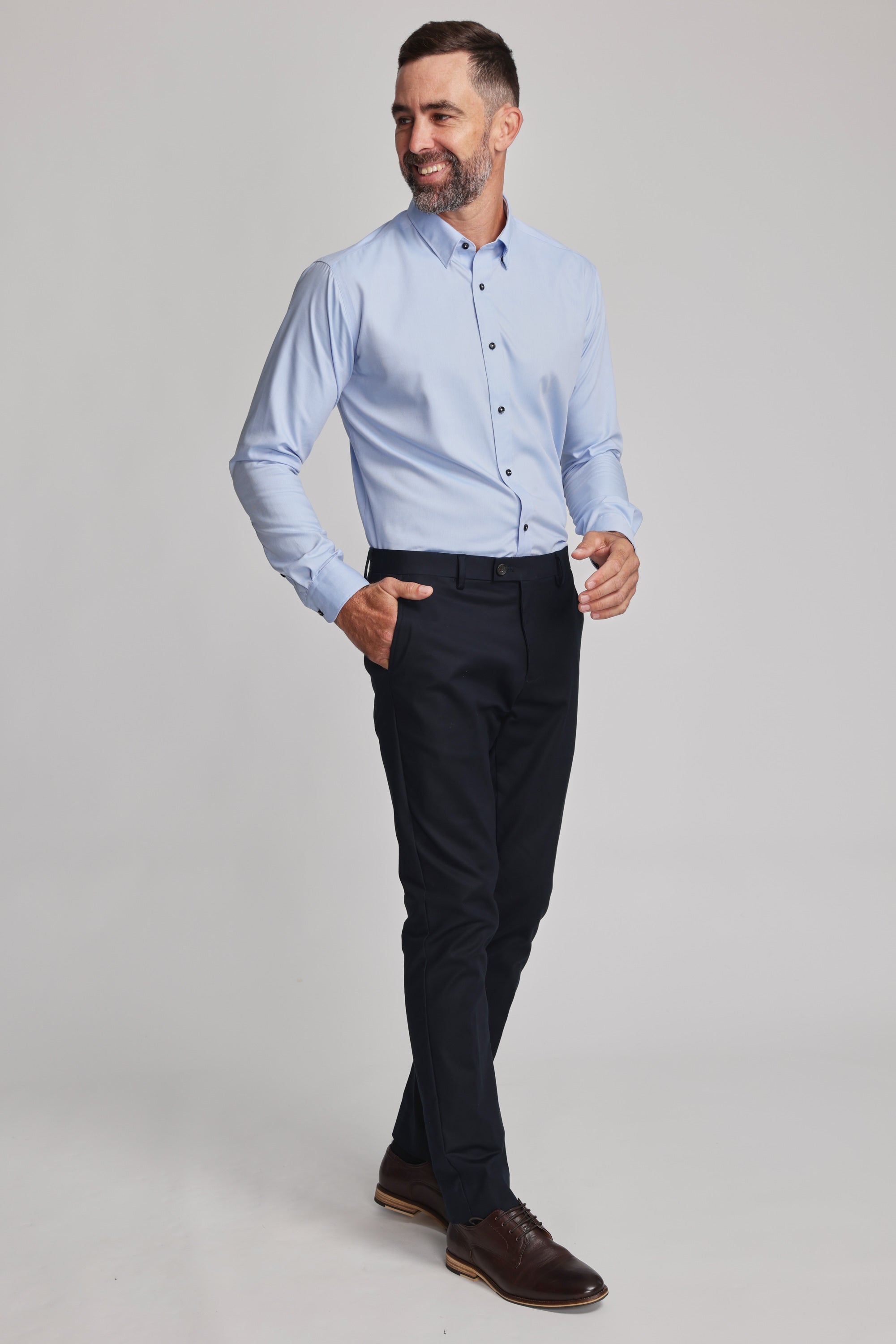 A man with short dark hair and a beard smiles as he walks in a Barkers Verona Shirt—tailored fit, long sleeve, 100% cotton—paired with black trousers and brown dress shoes against a plain light background.