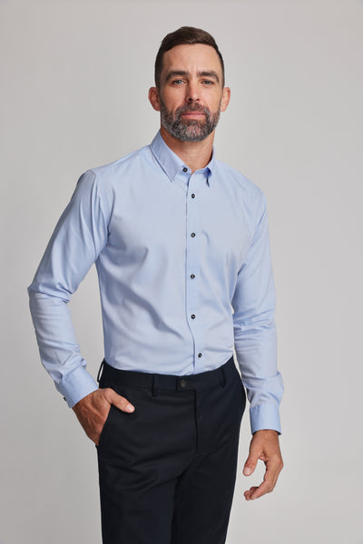 A man with short dark hair and a beard wears the Barkers Verona Shirt, a tailored fit light blue long sleeve business shirt, paired with dark pants. He stands against a plain light background with one hand in his pocket and a neutral expression.