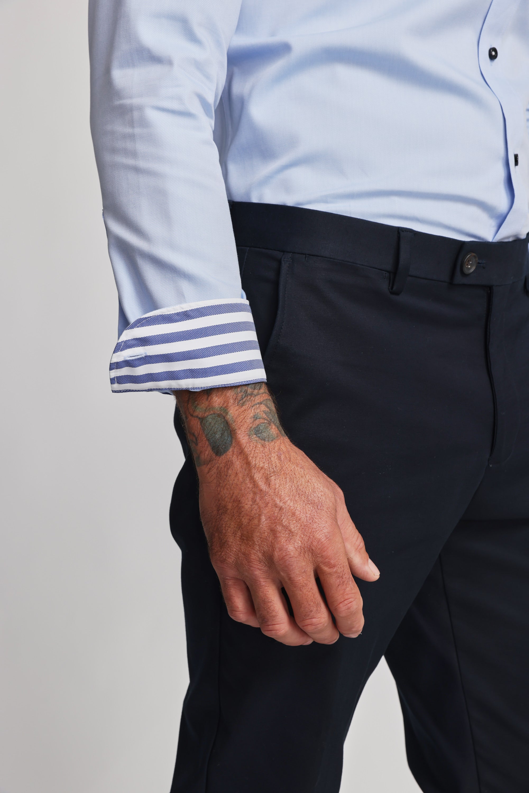 A person wears the Barkers Verona Shirt—100% cotton, tailored fit, light blue with striped inner cuffs—sleeve flipped to reveal stripes. They also wear dark pants and display a tattooed hand and forearm.