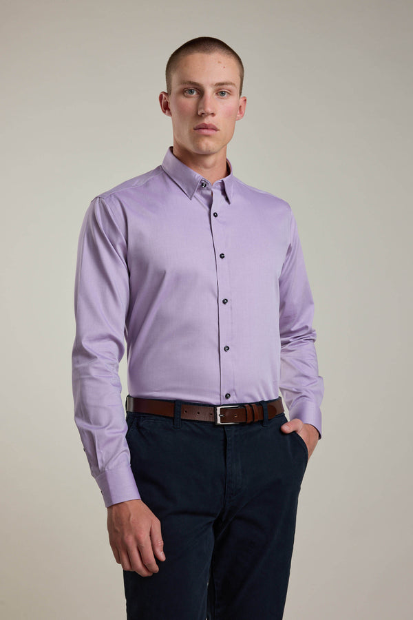 A young man faces forward wearing the Barkers Verona Shirt, a tailored fit, long sleeve business shirt in light purple, tucked into dark navy pants with a brown belt. One hand is in his pocket and he stands against a plain background.