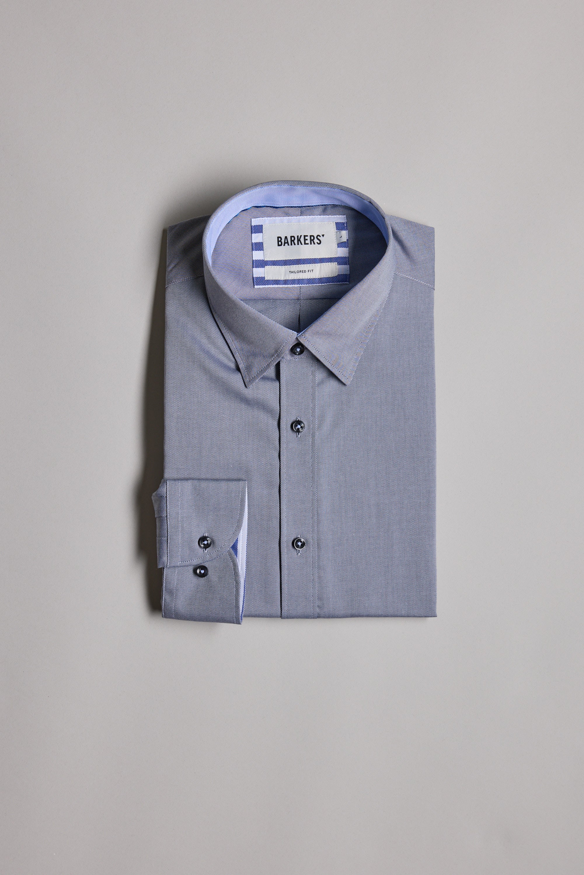 The Verona Shirt by Barkers is a neatly folded light blue long sleeve business shirt with dark buttons and striped inner collar fabric, featuring the BARKERS label, displayed on a plain grey background.