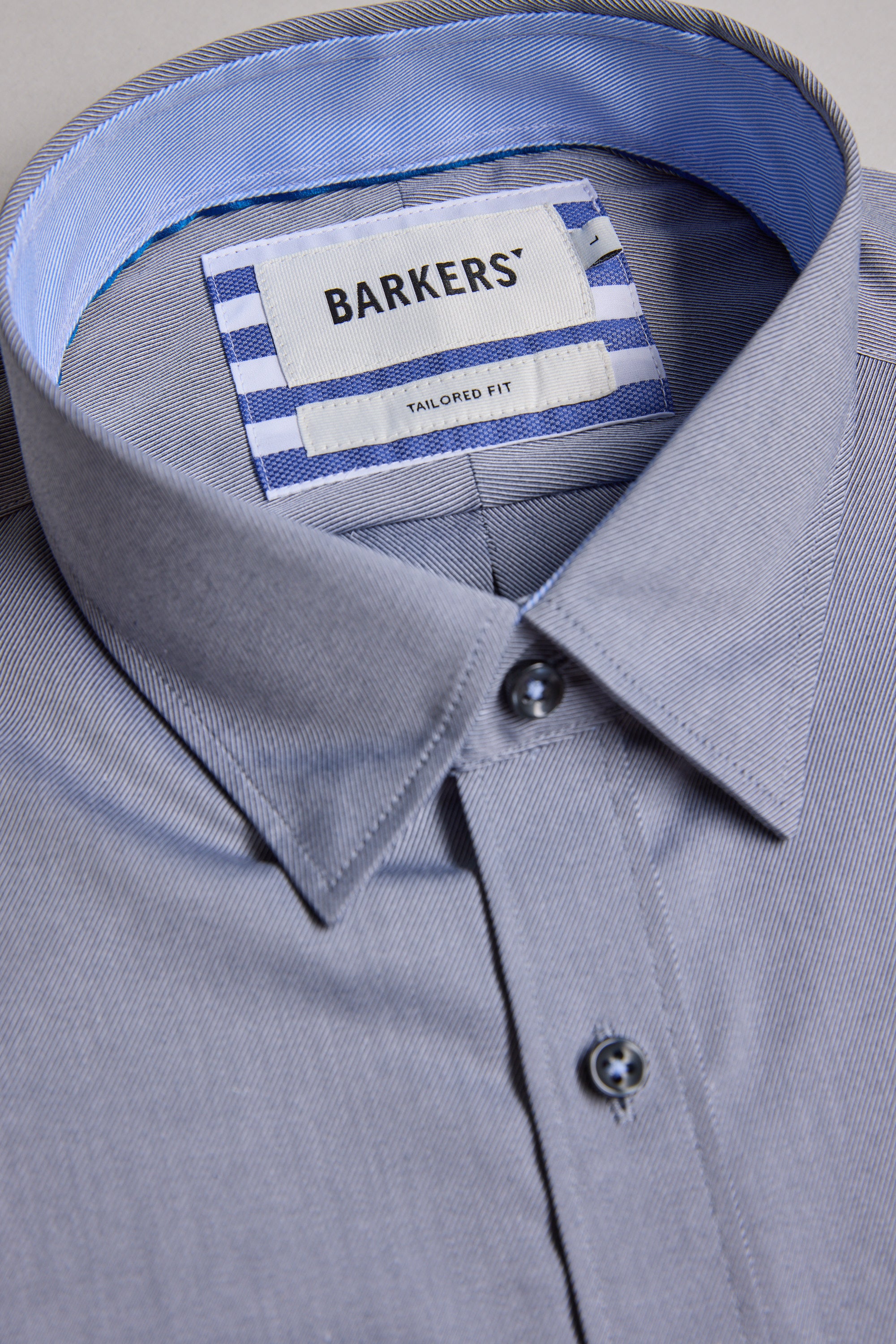 The Barkers Verona Shirt is a neatly folded light grey long sleeve business shirt with a pointed collar, blue buttons, and an inner label reading "Barkers Tailored Fit" on a white and blue striped background.