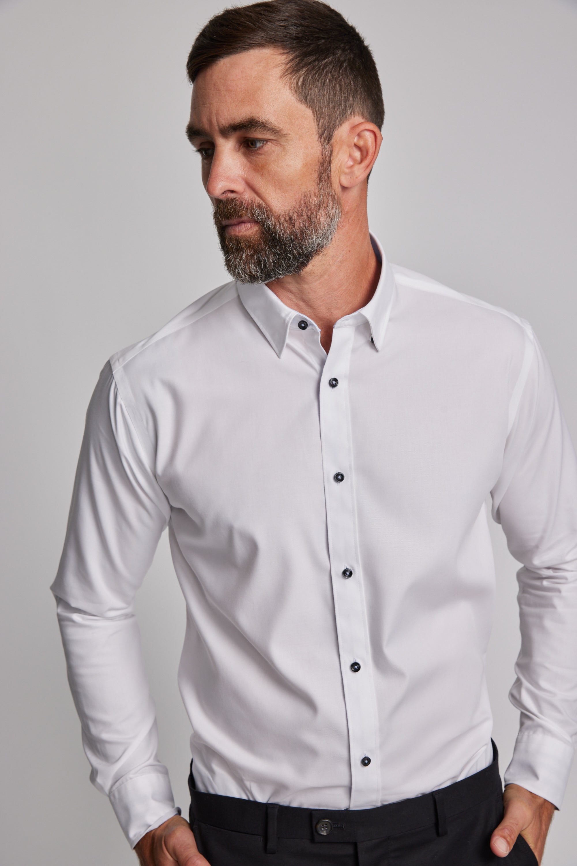 A man with short dark hair and a beard wears the Barkers Verona Shirt, a tailored fit, long sleeve 100% cotton business shirt, paired with dark pants. He stands against a plain light background, looking to his left.