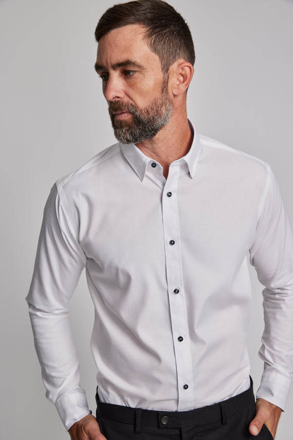A man with short dark hair and a beard wears the Barkers Verona Shirt, a tailored fit, long sleeve 100% cotton business shirt, paired with dark pants. He stands against a plain light background, looking to his left.