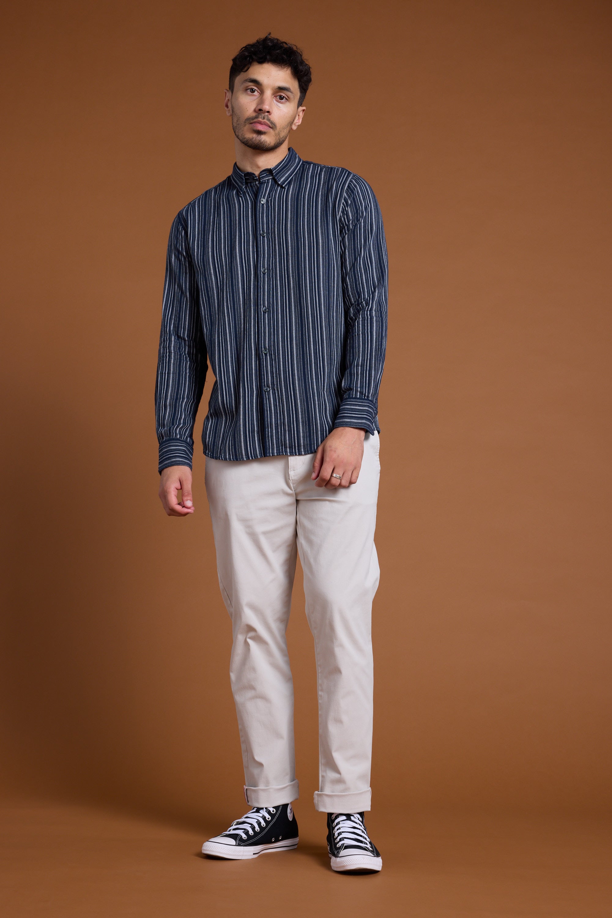 A man stands against a plain brown background, wearing Barkers' Scott Stripe Shirt in navy and white, made from responsibly sourced cotton, paired with light beige pants and black high-top sneakers. He looks at the camera with a neutral expression.
