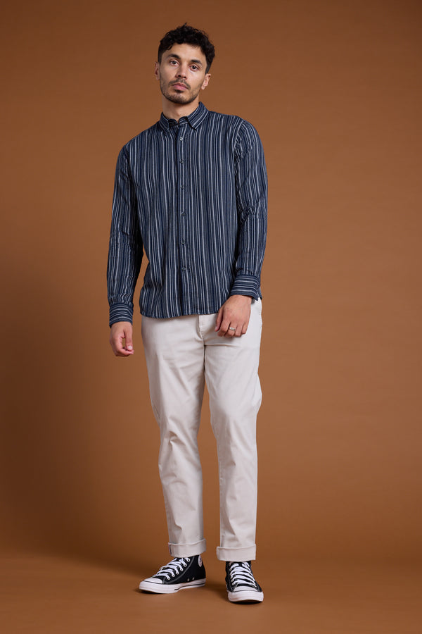 A man stands against a plain brown background, wearing Barkers' Scott Stripe Shirt in navy and white, made from responsibly sourced cotton, paired with light beige pants and black high-top sneakers. He looks at the camera with a neutral expression.