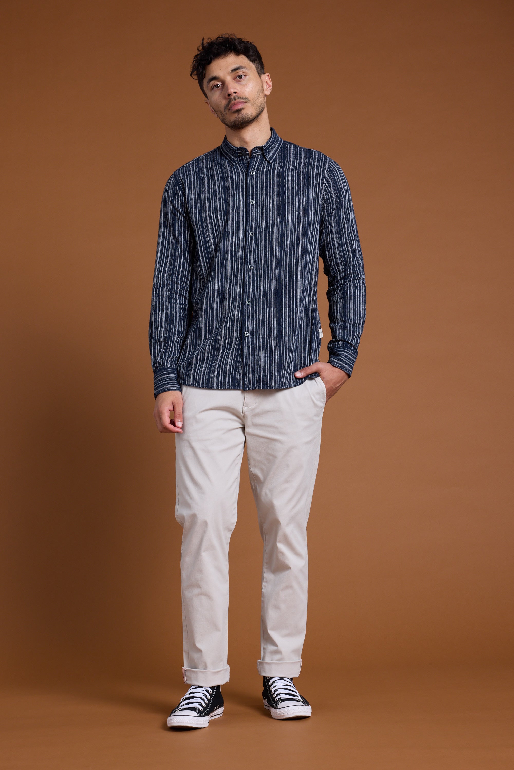 A man stands against a brown background wearing the Barkers Scott Stripe Shirt in a tailored fit, made from responsibly sourced cotton, paired with beige pants and black and white sneakers. One hand is in his pocket as he looks to the side.