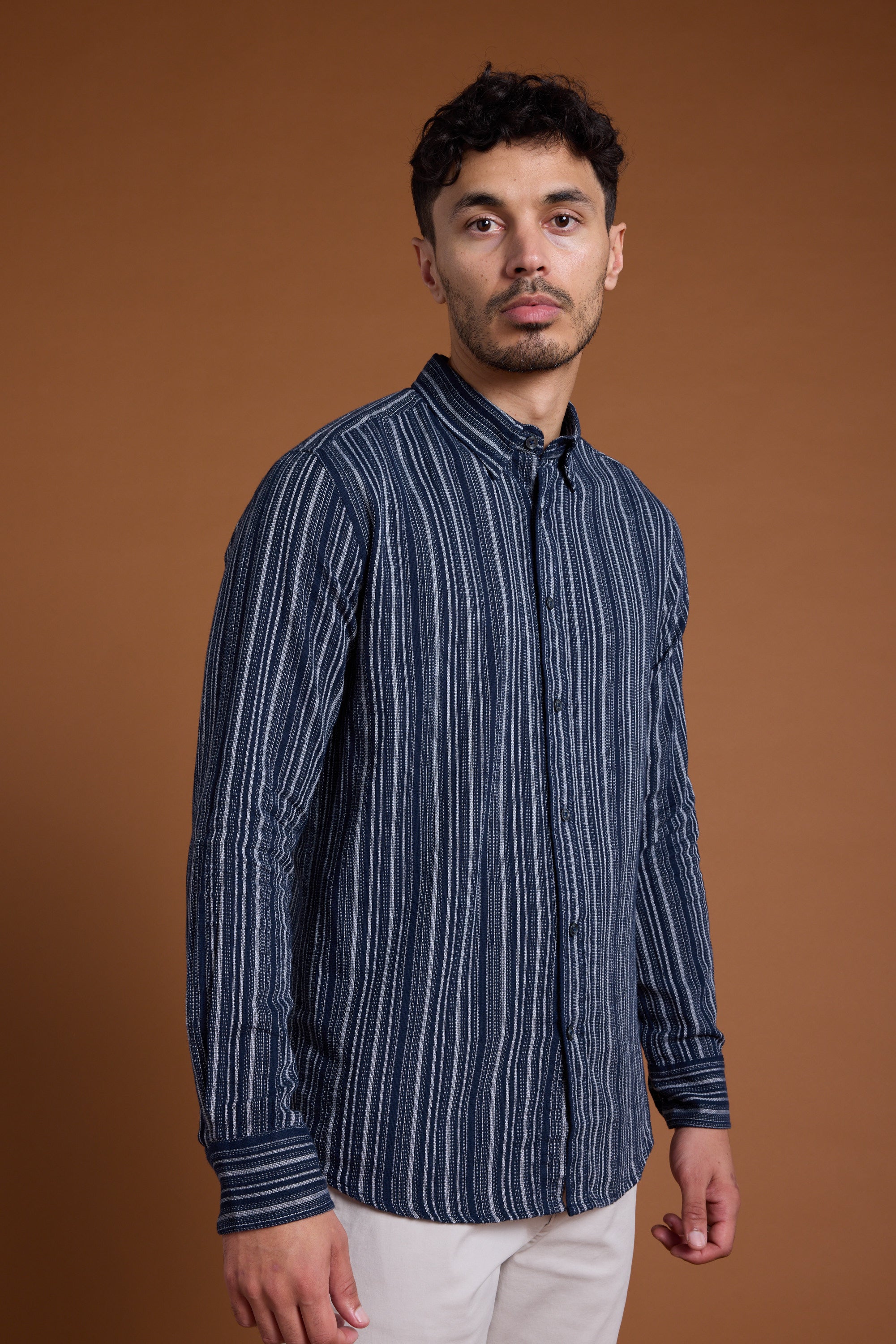 A man with short dark hair and a trimmed beard wears the Barkers Scott Stripe Shirt—a navy and white tailored fit shirt made from responsibly sourced cotton—paired with light pants, standing against a plain brown background.