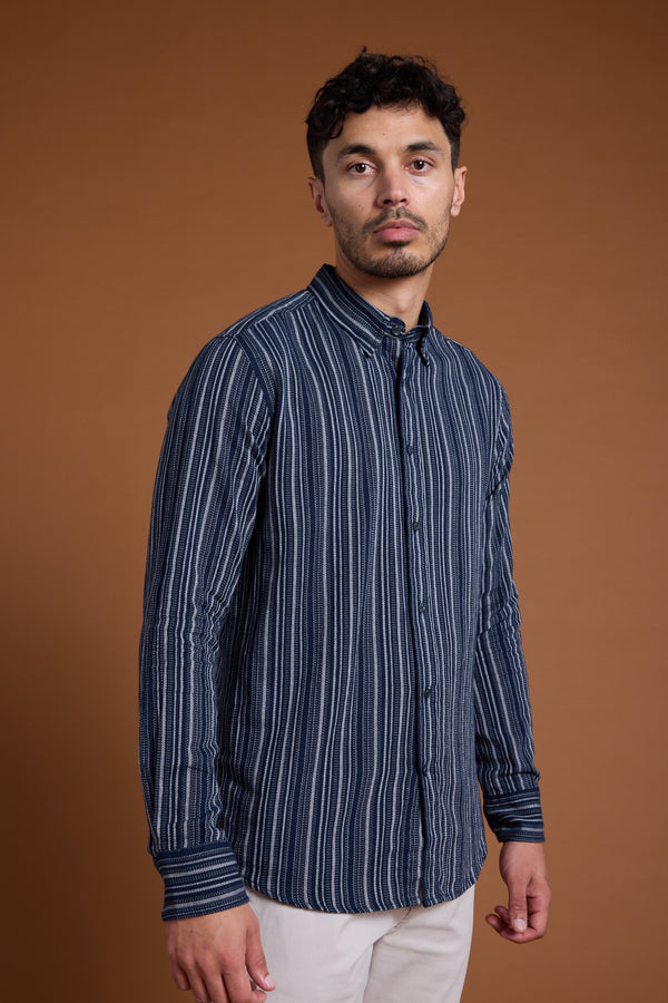 A man with short dark hair and a trimmed beard wears the Barkers Scott Stripe Shirt—a navy and white tailored fit shirt made from responsibly sourced cotton—paired with light pants, standing against a plain brown background.