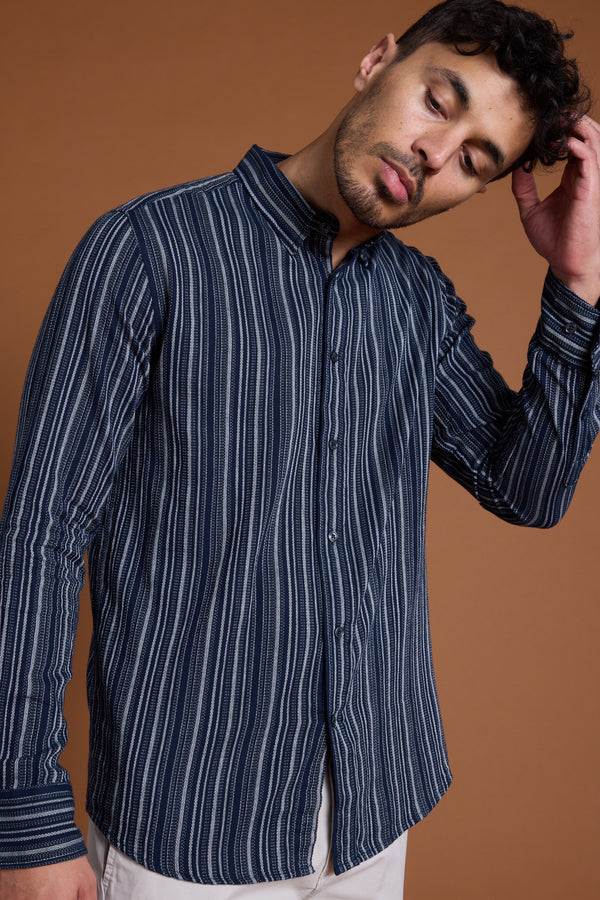 A man wears the Barkers Scott Stripe Shirt in navy and white, made from responsibly sourced cotton, with light pants. He stands against a brown background, tilting his head and touching his hair with one hand.
