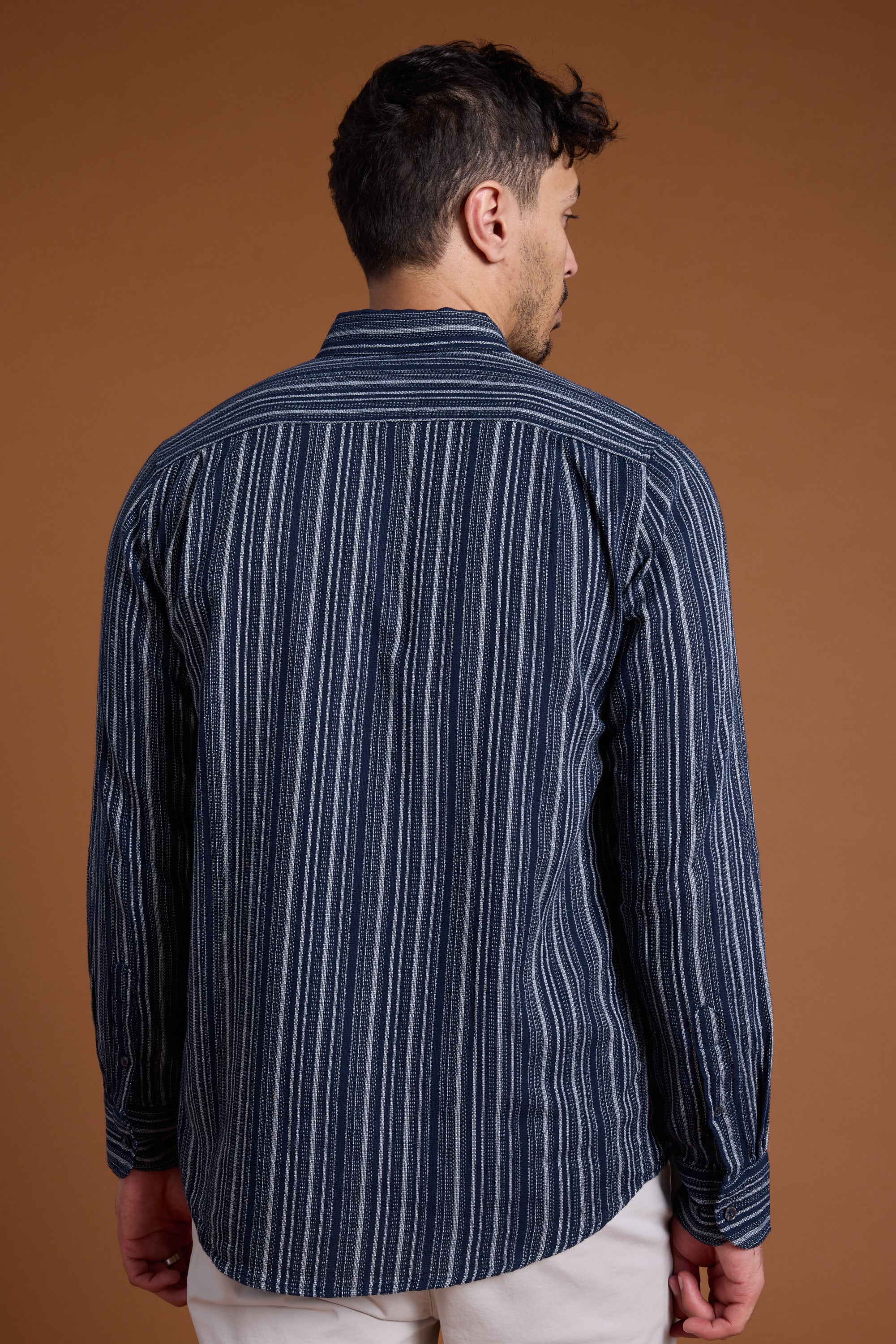 A man stands facing away against a plain brown background, wearing Barkers' Scott Stripe Shirt—long-sleeved, dark blue with vertical white stripes, made from responsibly sourced cotton—and light-colored pants.