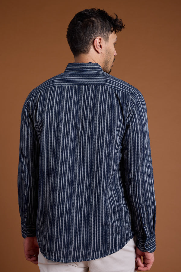 A man stands facing away against a plain brown background, wearing Barkers' Scott Stripe Shirt—long-sleeved, dark blue with vertical white stripes, made from responsibly sourced cotton—and light-colored pants.