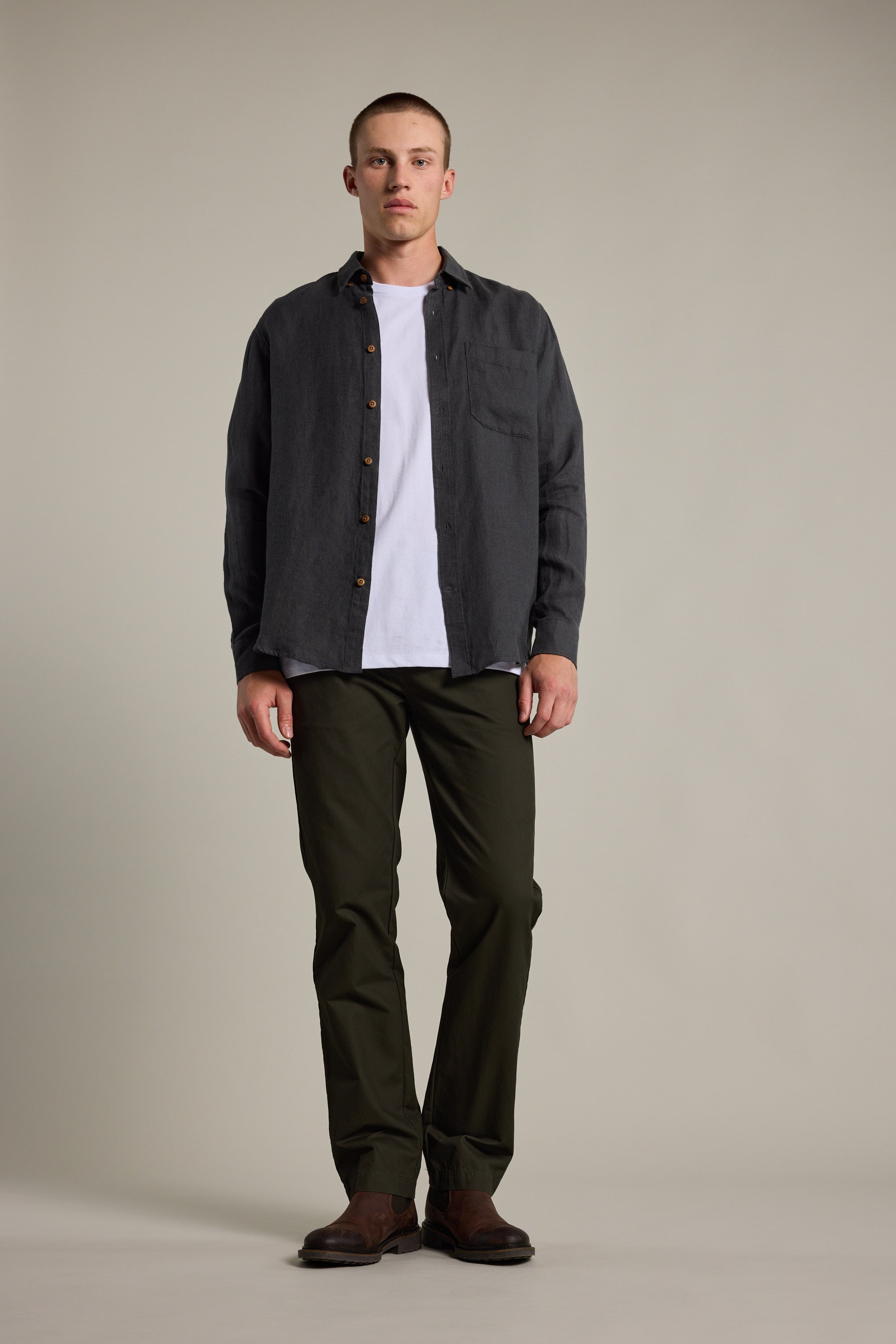 A man faces forward against a plain background, wearing the Barkers Delano Linen Shirt in dark gray over a white tee, paired with olive green pants and brown shoes.