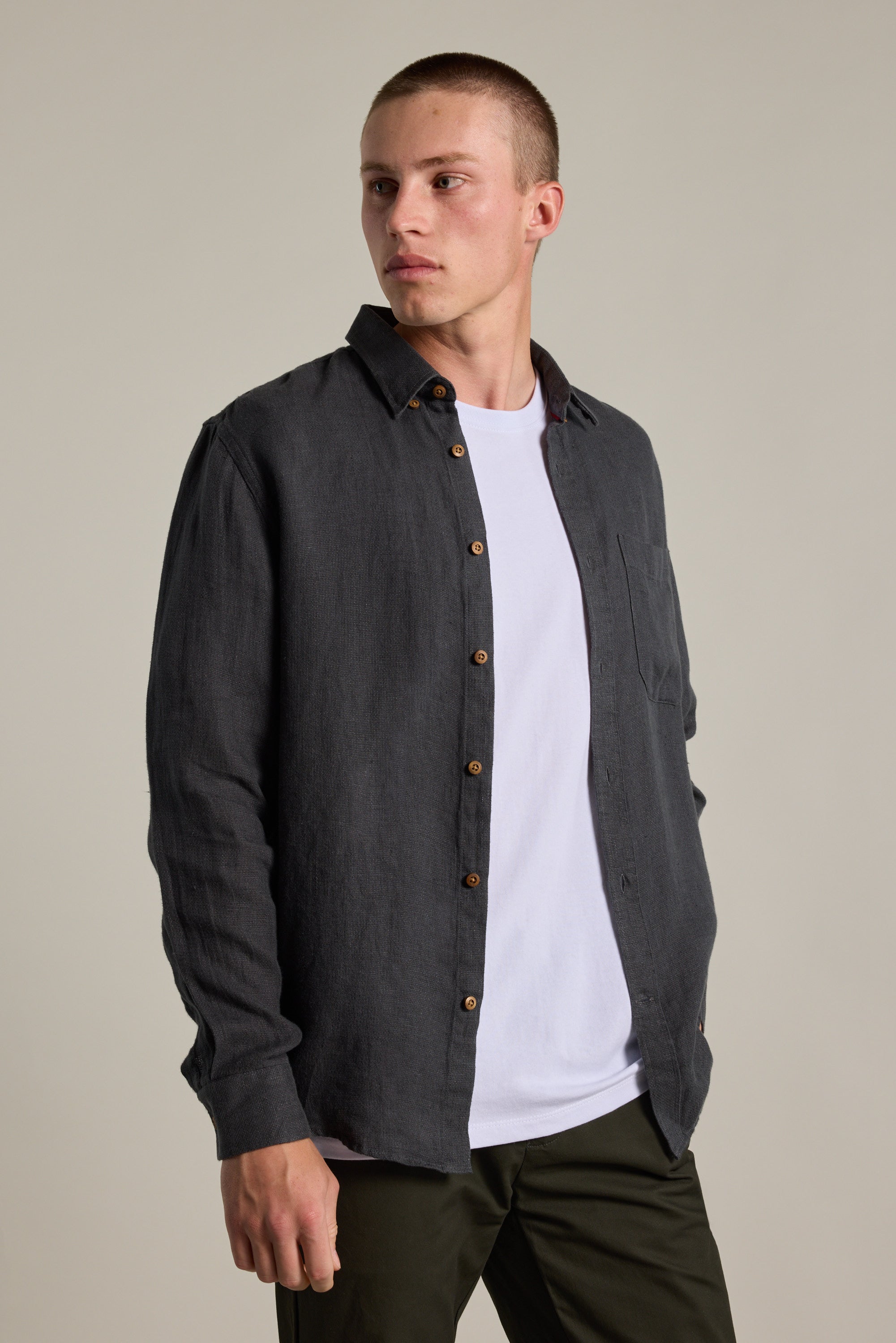 A person wearing the Barkers Delano Linen Shirt in a classic dark shade over a white tee and dark pants stands slightly sideways, looking left against a plain light background.