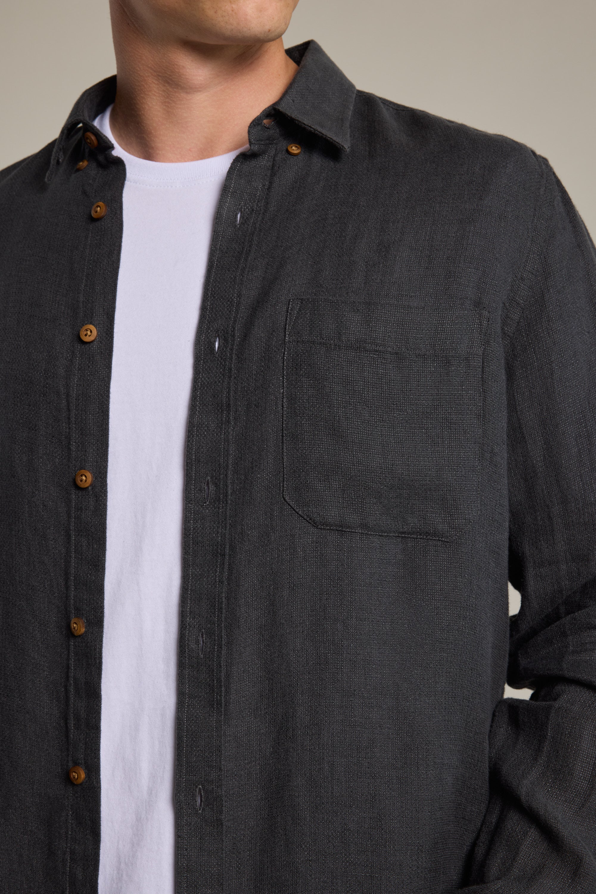 A person is shown from shoulders to waist with their left hand in a pocket, wearing the Barkers Delano Linen Shirt in classic fit dark gray hammered linen with brown buttons, layered over a white t-shirt.