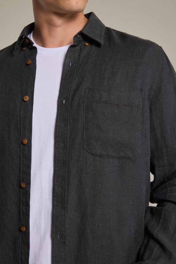 A person is shown from shoulders to waist with their left hand in a pocket, wearing the Barkers Delano Linen Shirt in classic fit dark gray hammered linen with brown buttons, layered over a white t-shirt.