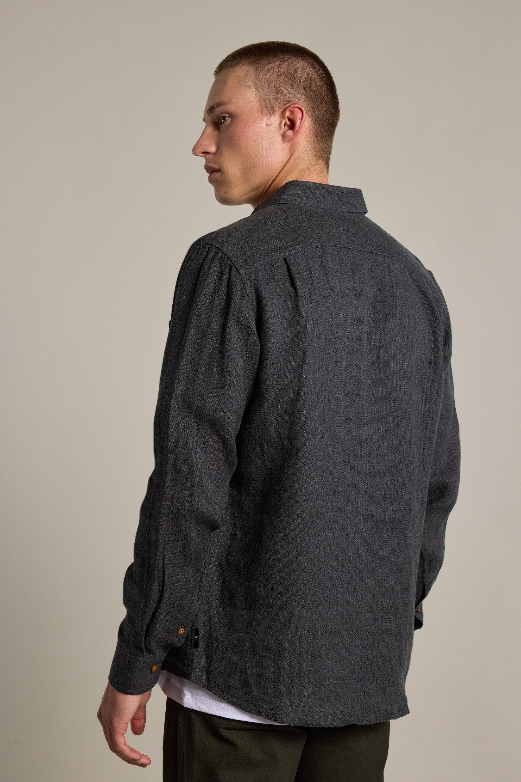 A young man with short hair models the Barkers Delano Linen Shirt in a classic fit, layered over a white tee, as he faces away and looks over his left shoulder against a plain light background.
