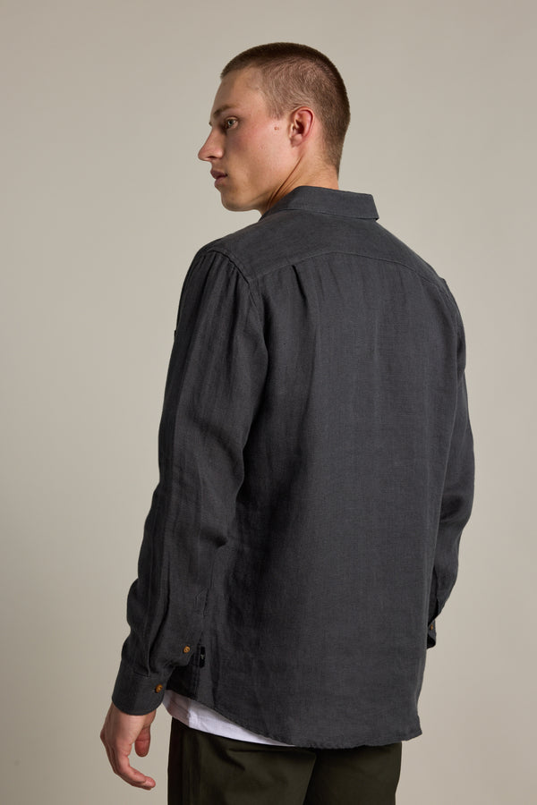 A young man with short hair models the Barkers Delano Linen Shirt in a classic fit, layered over a white tee, as he faces away and looks over his left shoulder against a plain light background.