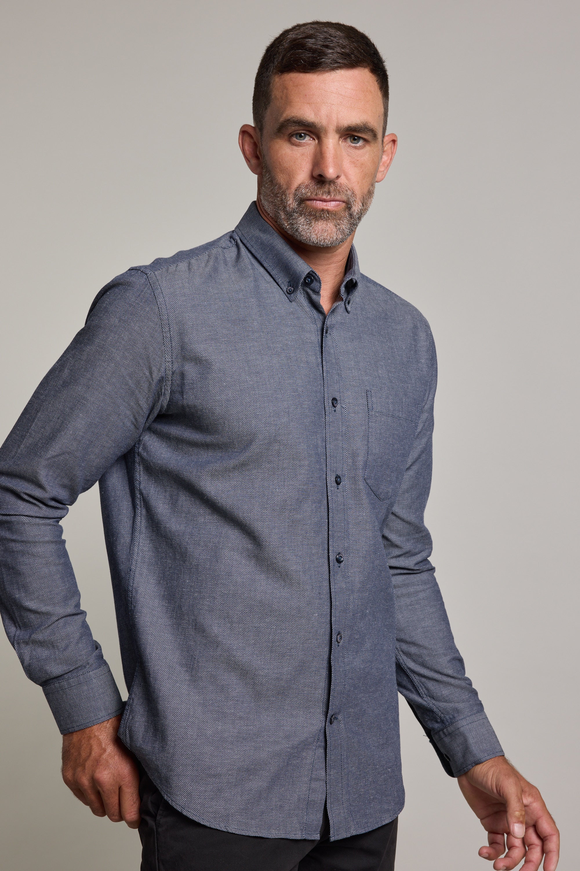 A man with short hair and a beard wears the Barkers Tyson Texture Shirt, a long-sleeved, blue-gray linen-cotton blend button-up, paired with dark pants, standing against a plain light background and looking confidently at the camera.