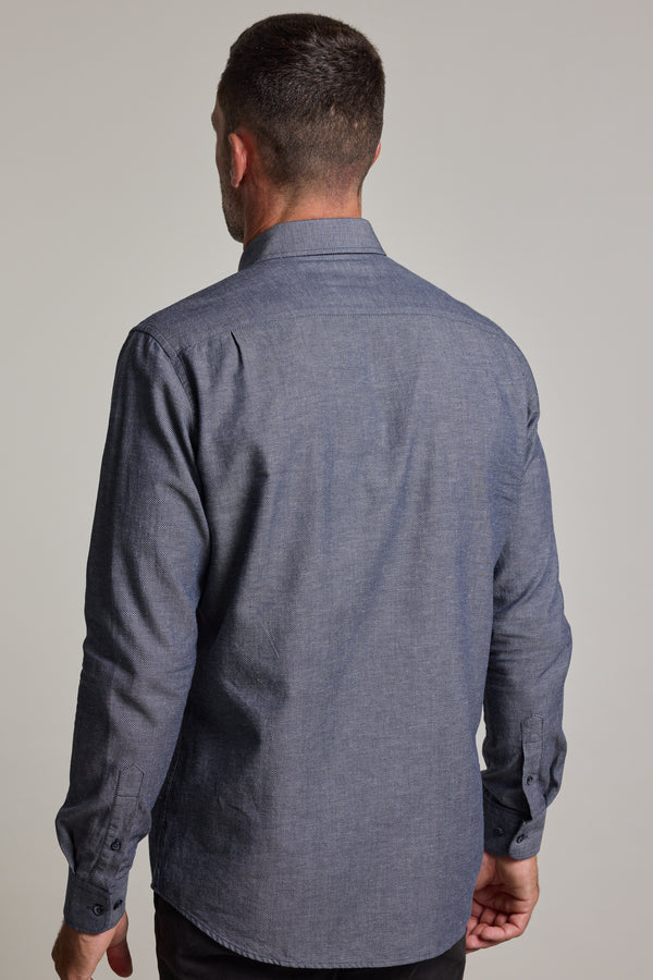 A man faces away from the camera, wearing the Barkers Tyson Texture Shirt—a long-sleeved, blue textured, tailored-fit button-up—paired with dark pants. The background is plain and light-colored.