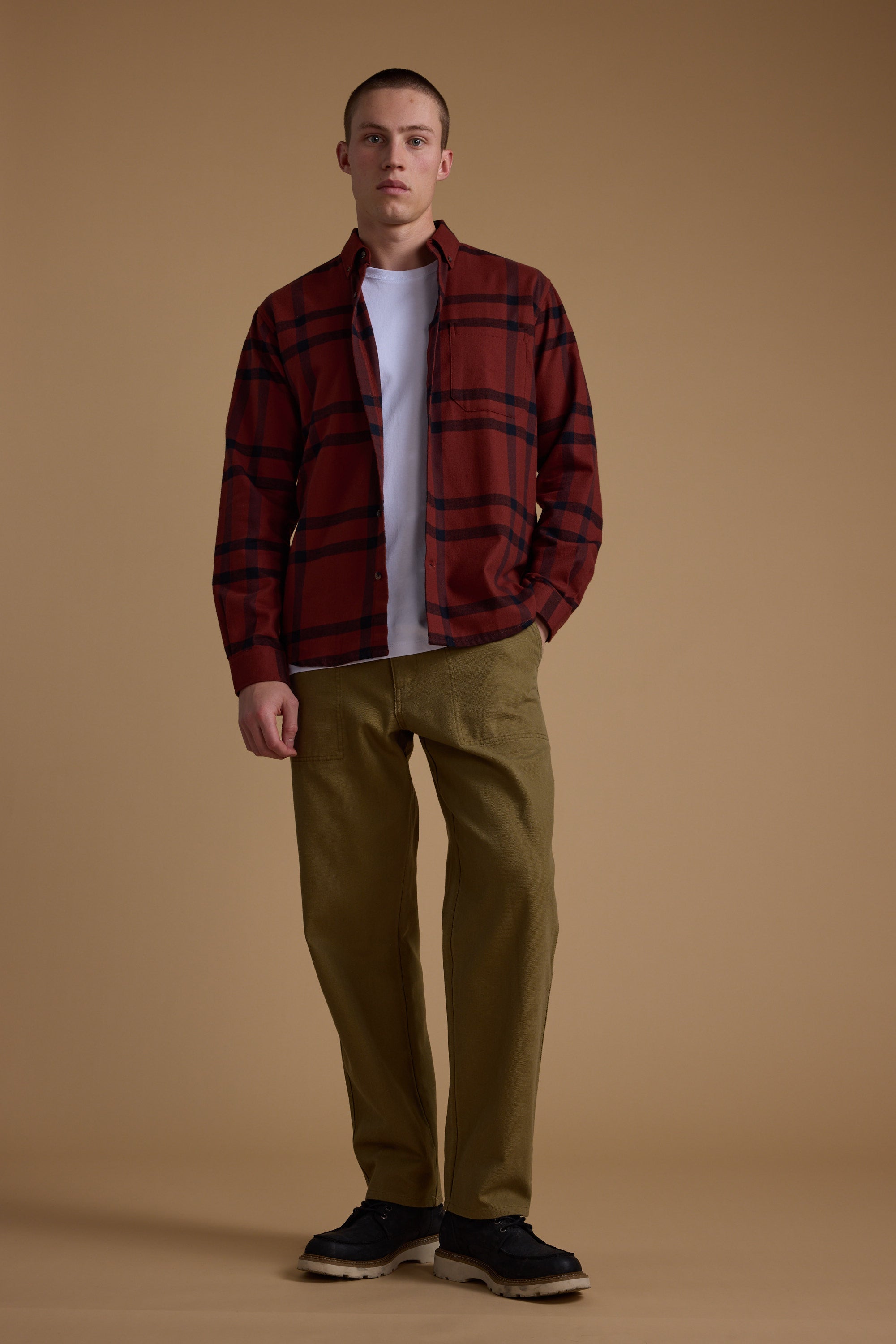 A young man stands against a brown background, wearing the Barkers Durness Check Shirt in red flannel over a white t-shirt, with olive green pants and black shoes. He faces forward with one hand in his pocket.