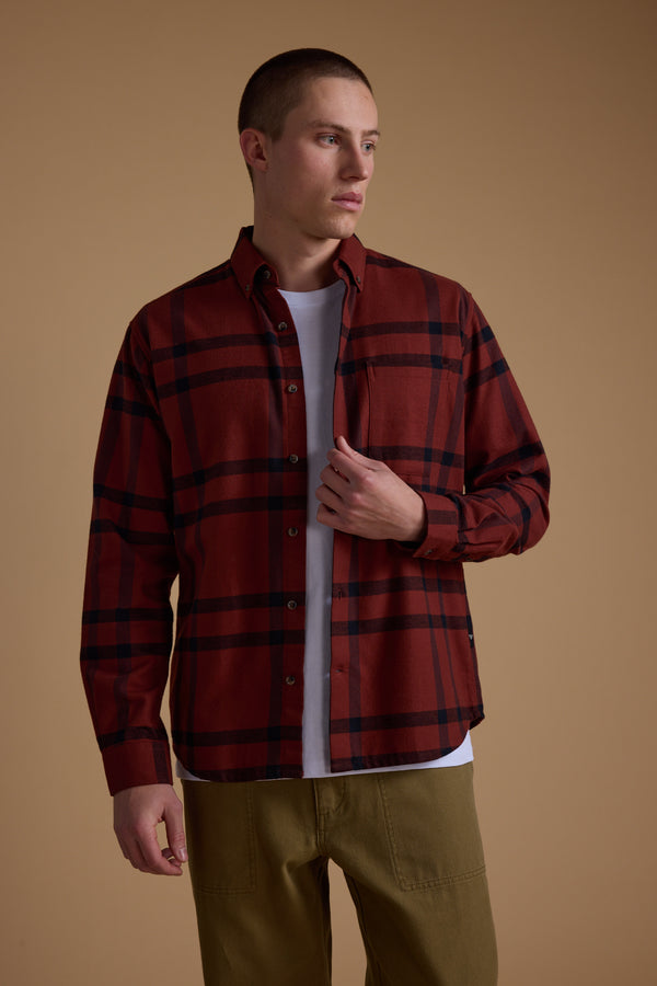 A person wears the Barkers Durness Check Shirt—a red flannel made from responsibly sourced cotton—over a white tee with olive green pants, standing against a tan background and looking to the side with a neutral expression.