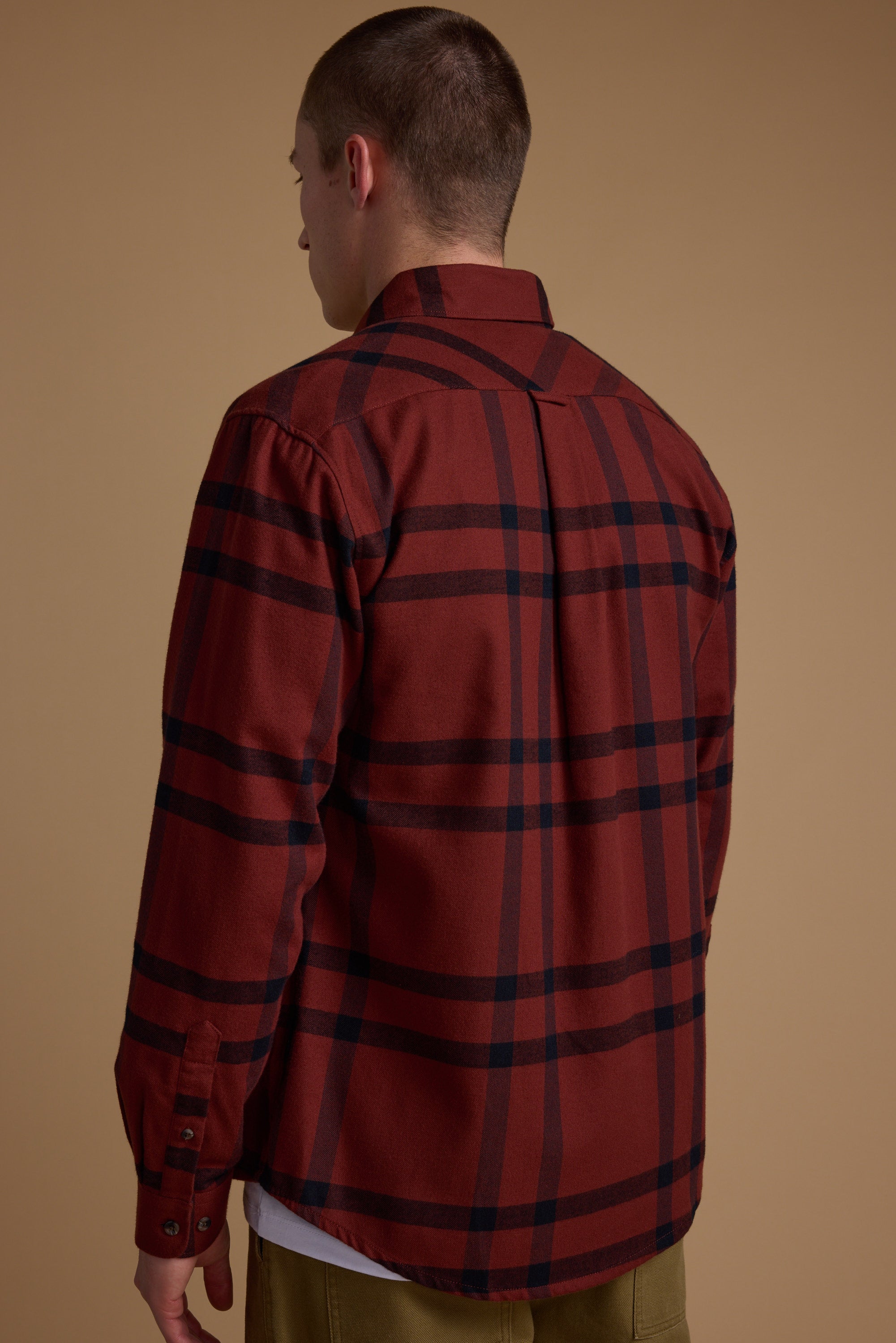 A person stands with their back to the camera, wearing a red Durness Check Shirt by Barkers over a white shirt and olive pants, against a plain brown background. The flannel shirt is made from responsibly sourced cotton.