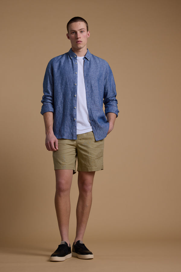 A person stands before a tan backdrop wearing Barkers' Jasper Linen Weekend Short, a blue button-up shirt over a white tee, and black sneakers, looking directly at the camera with a neutral expression.