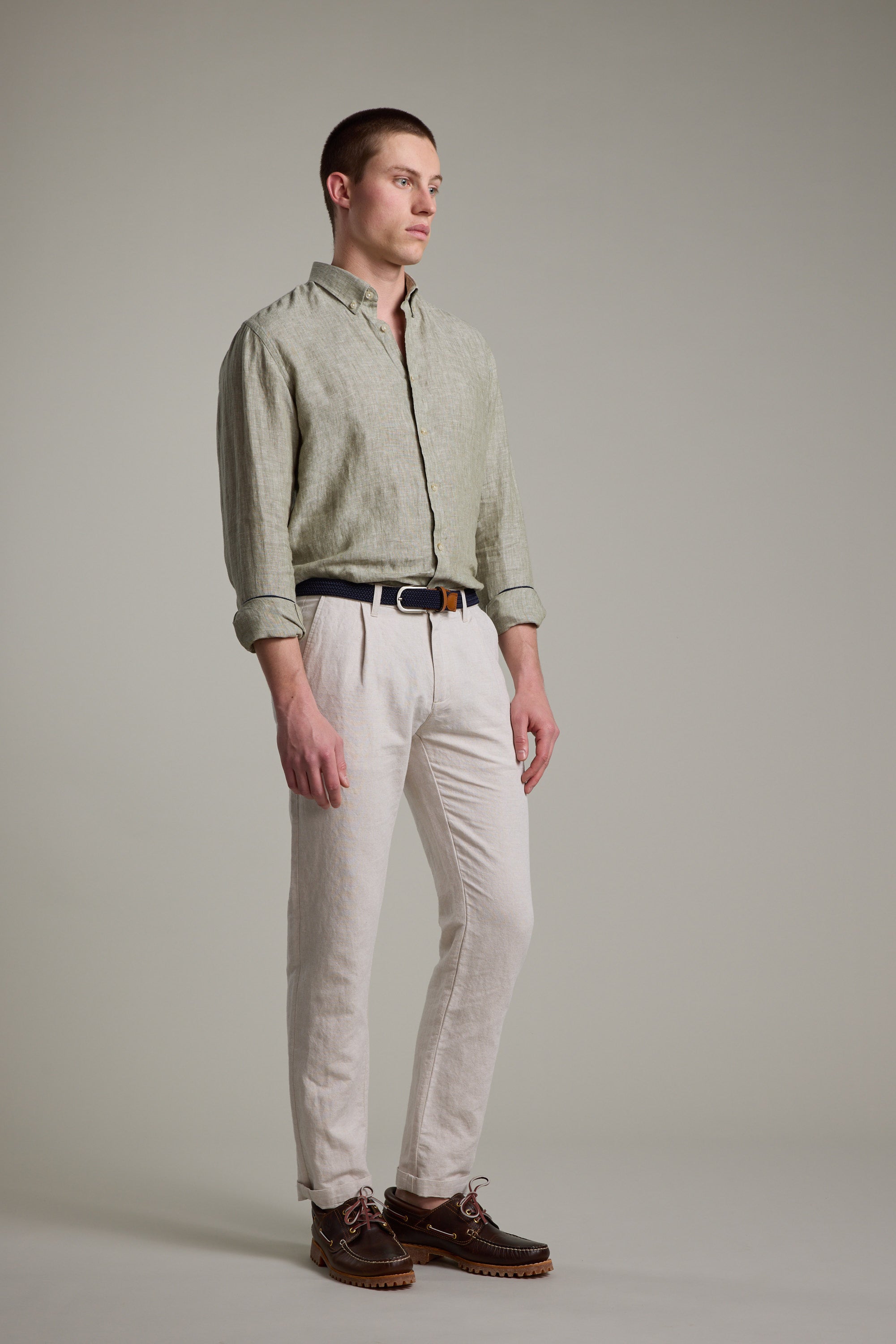 A man with short hair stands against a plain background in Barkers’ Dorset Linen Shirt, light green with rolled sleeves, tailored fit, paired with beige pants, a black belt, and brown shoes.