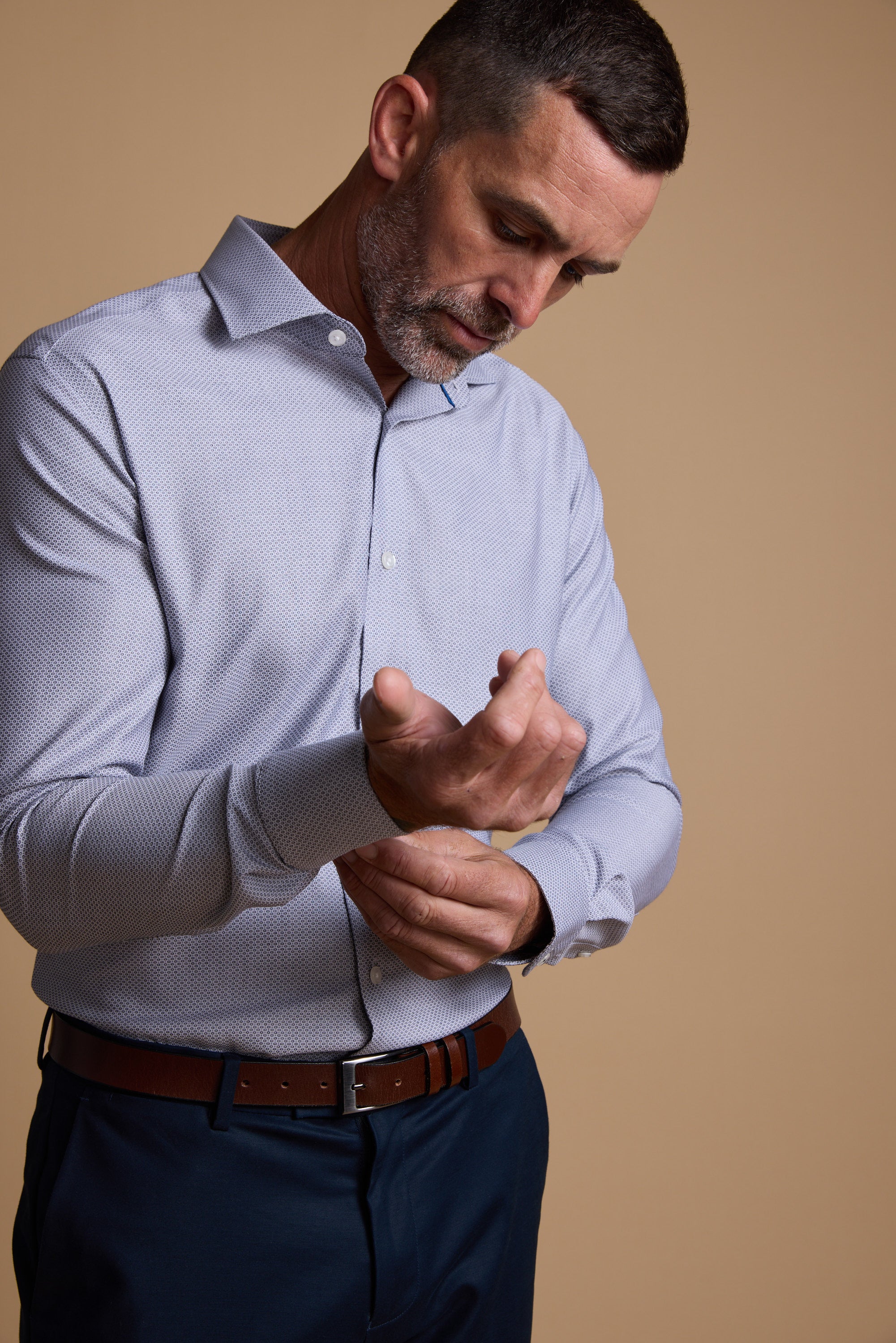 A man with short dark hair and a beard buttons the cuff of his light blue Barkers Eaton Jacquard Business Shirt, paired with dark pants and a brown belt, standing against a plain beige background.