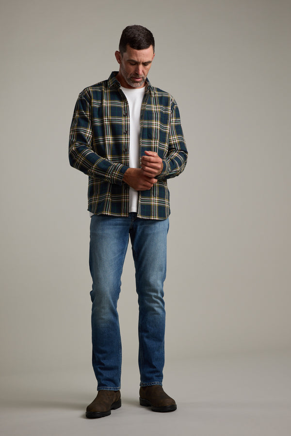 A man stands against a plain background, wearing the Barkers Brennan Check Overshirt over a white tee, blue jeans, and brown boots. He looks down as he adjusts his shirt sleeve with both hands.