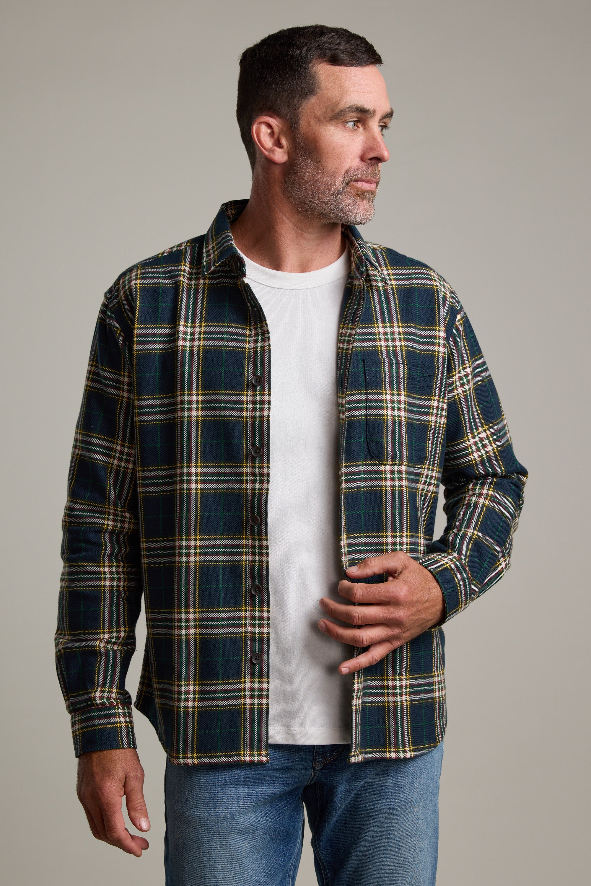 A man with short dark hair and a beard wears the Barkers Brennan Check Overshirt in green and yellow plaid over a white t-shirt and blue jeans, standing against a plain background and looking to his left.