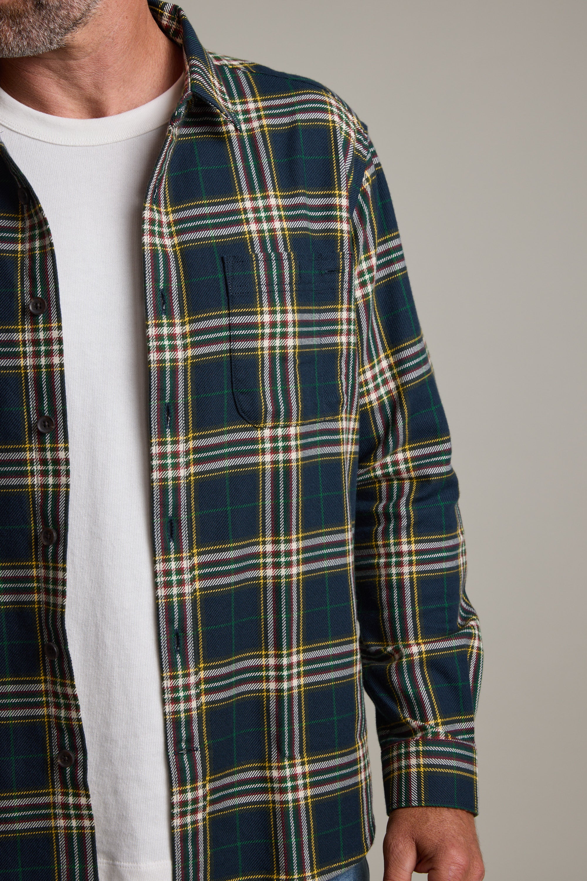 A person wears a white T-shirt under an open, green and yellow Barkers Brennan Check Overshirt. Only the lower half of the face, torso, and right arm are visible against a plain background. Made from responsibly sourced cotton.