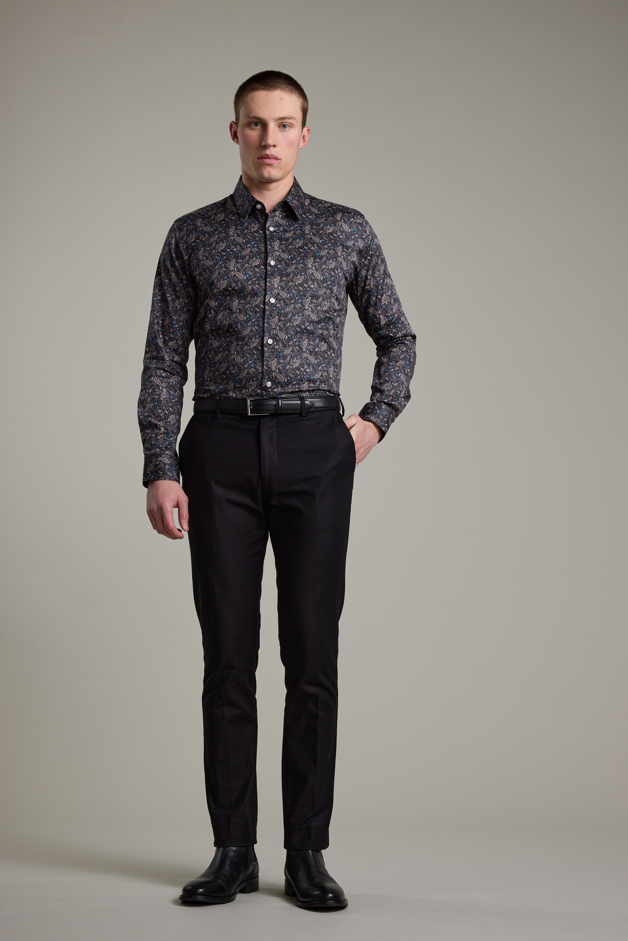 A man with short hair and a neutral expression wears the Barkers Hilton Print Shirt, featuring a dark floral design, tucked into black dress pants with a belt and black shoes. He stands against a plain background with one hand in his pocket.