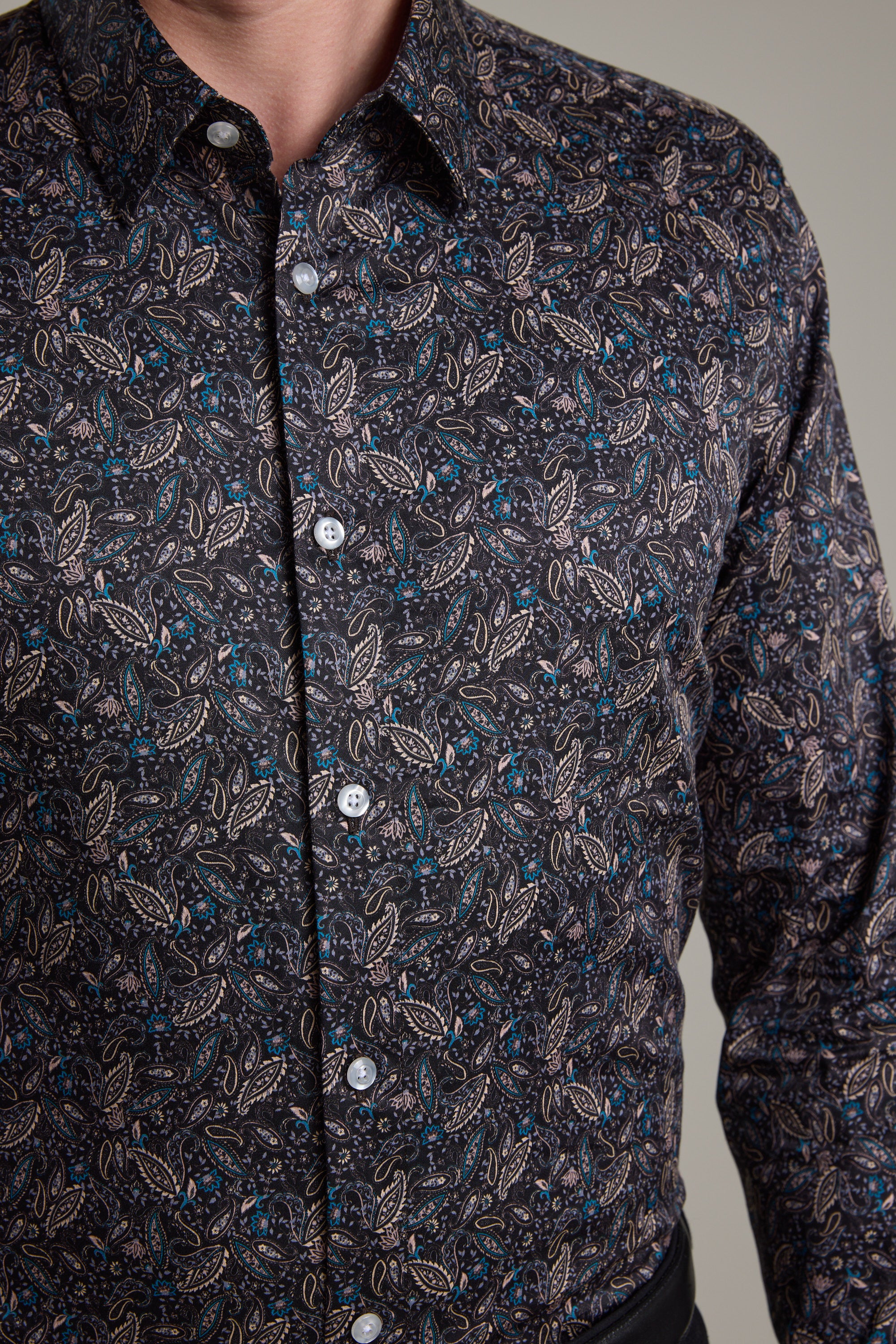 A person wears the Barkers Hilton Print Shirt, featuring a detailed paisley pattern in light brown, blue, and cream. Made from responsibly sourced cotton, this tailored fit dark button-up is styled with black pants.