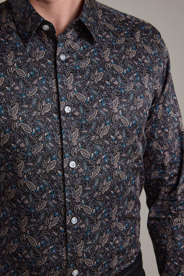 A person wears the Barkers Hilton Print Shirt, featuring a detailed paisley pattern in light brown, blue, and cream. Made from responsibly sourced cotton, this tailored fit dark button-up is styled with black pants.