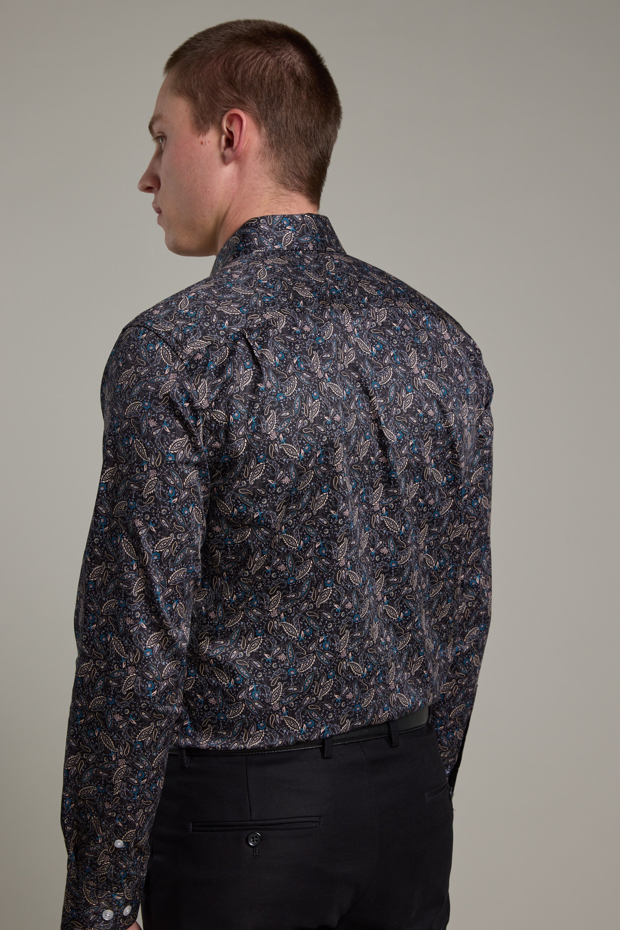 A man is seen from behind wearing the Barkers Hilton Print Shirt, a long-sleeved, dark floral design paired with black pants. The tailored fit stands out against a plain, neutral background.