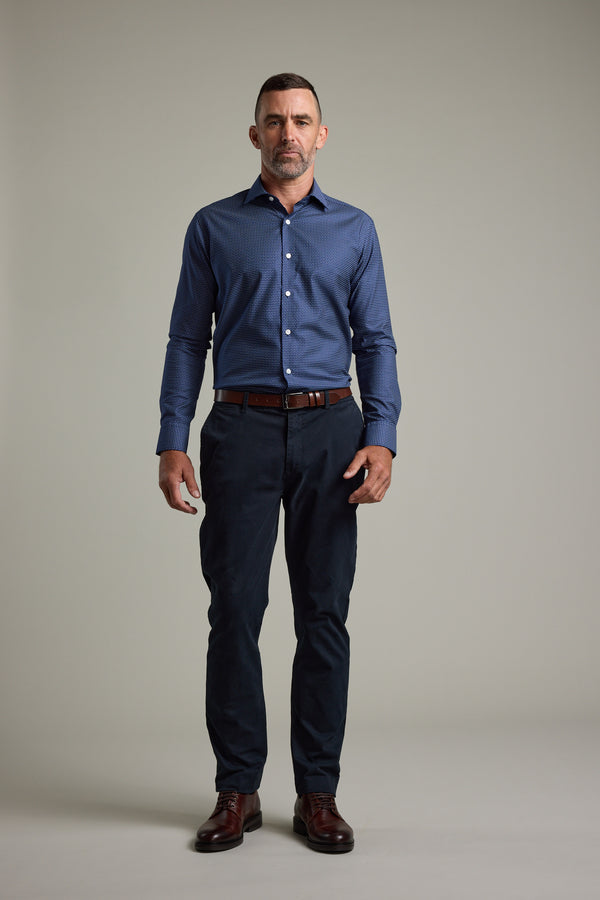 A man with short hair and a trimmed beard stands against a plain background, wearing the Barkers Maiden Paisley Business Shirt, dark trousers, a brown belt, and brown shoes. He faces the camera with a neutral expression.