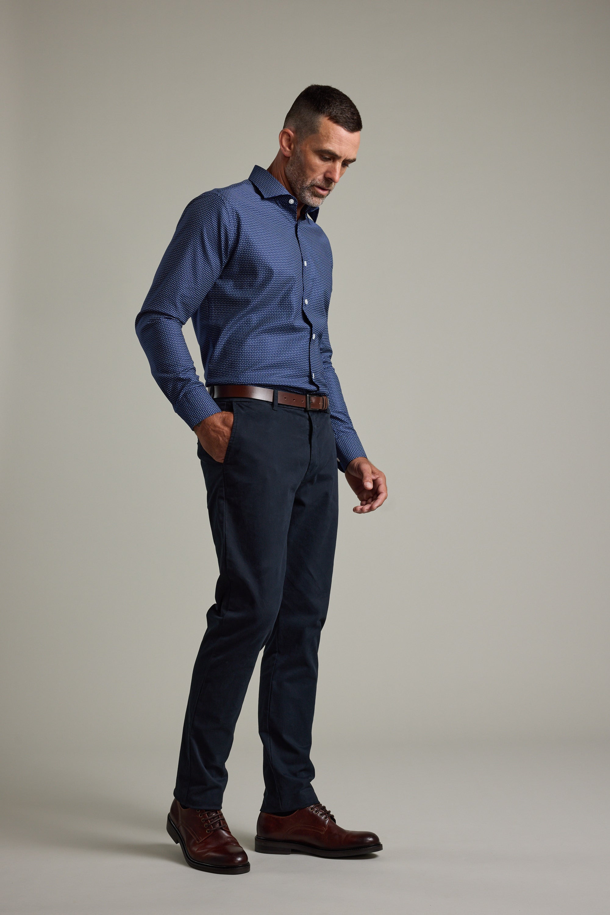 A man stands against a plain background wearing the Barkers Maiden Paisley Business Shirt, dark pants, a brown belt, and brown dress shoes. He looks down with one hand in his pocket.