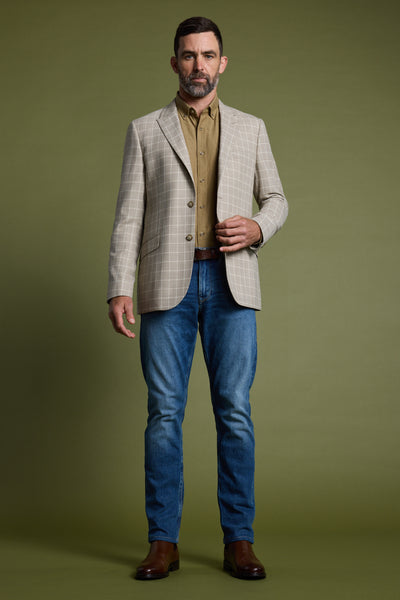 A man with short brown hair and a beard stands against an olive green background, wearing the Barkers Prestwick Check Blazer, a light brown shirt, blue jeans, and brown boots.