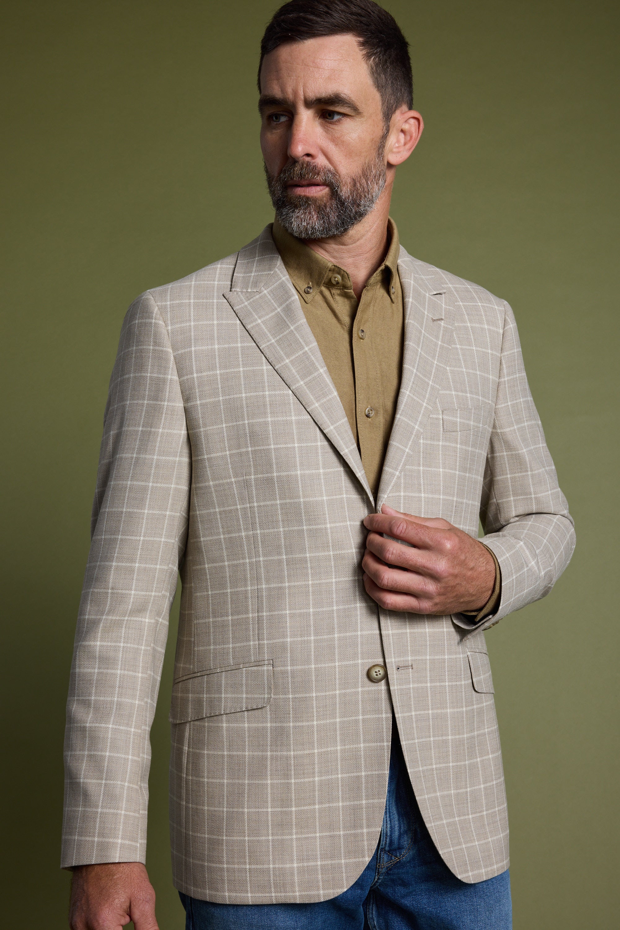 A man with short dark hair and a beard wears the Barkers Prestwick Check Blazer over a tan button-up shirt and blue jeans, standing against an olive green background while looking to the side.