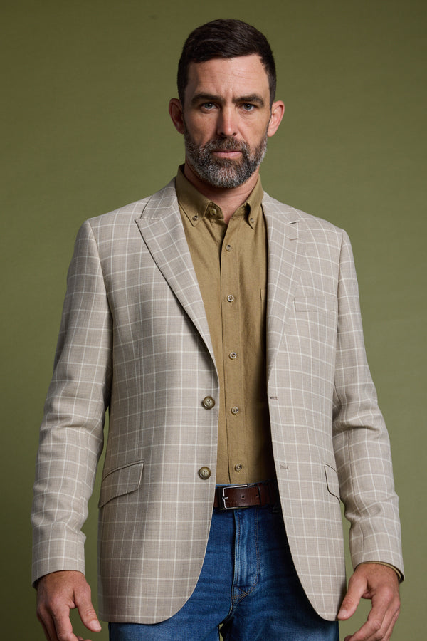 A man with short dark hair and a beard wears the Barkers Prestwick Check Blazer over a tan button-up shirt and blue jeans, standing against a plain olive green background.