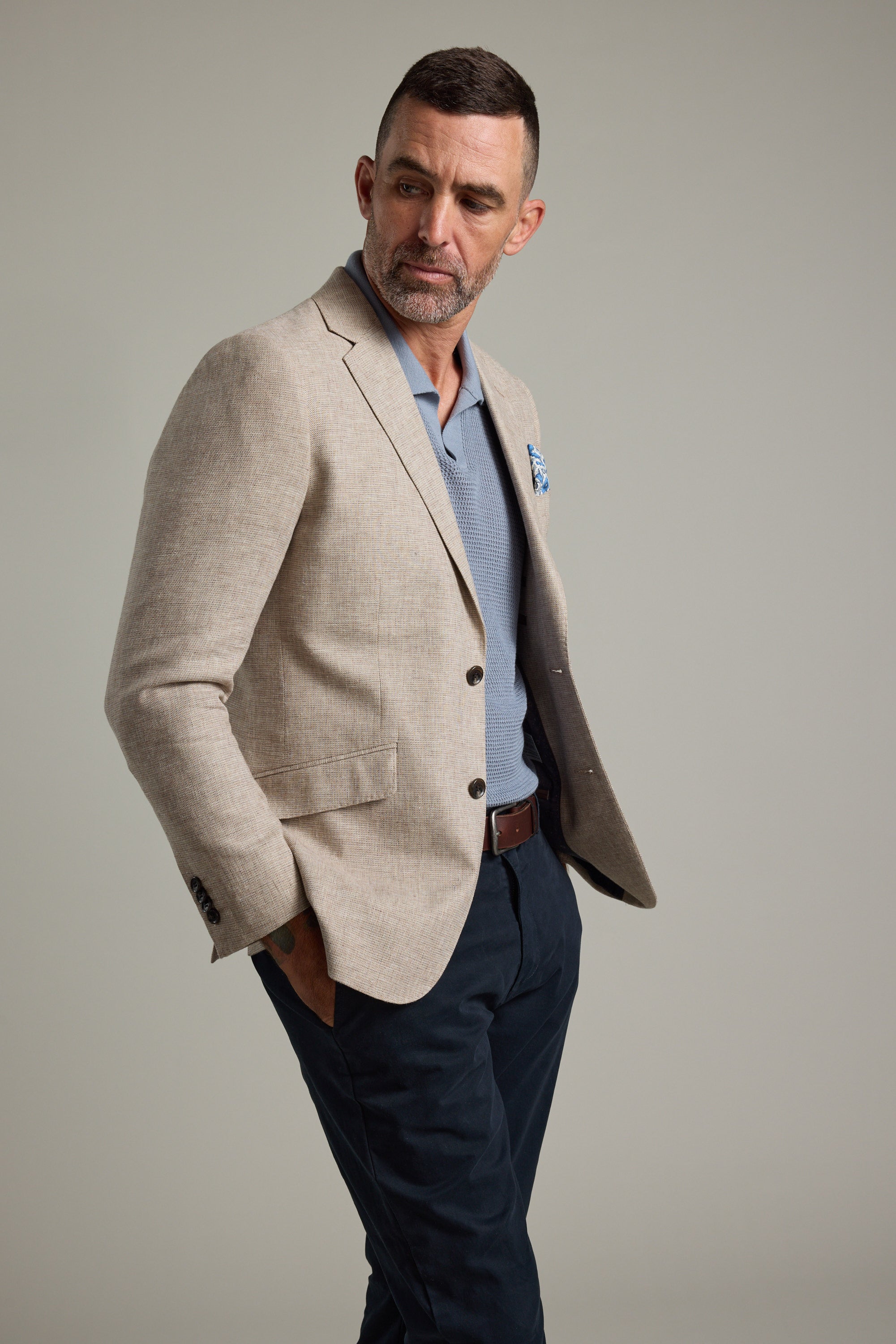 A man with short dark hair and a beard wears the Welland Texture Blazer by Barkers over a light blue shirt, dark pants, and a brown belt, standing with one hand in his pocket and looking to his left against a plain background.