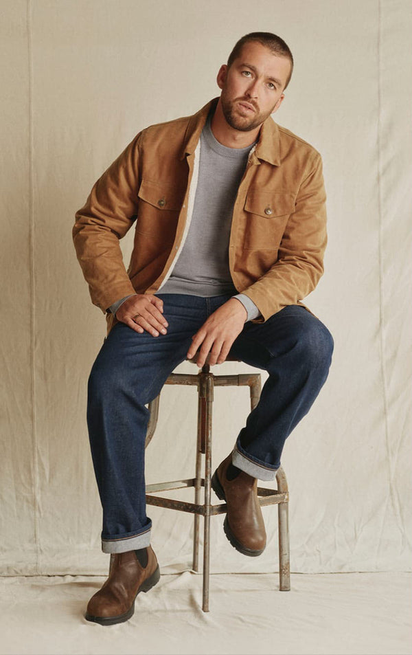 Man in tan jacket, grey shirt, blue jeans, and boots sits NZ-style on a stool against a plain backdrop.