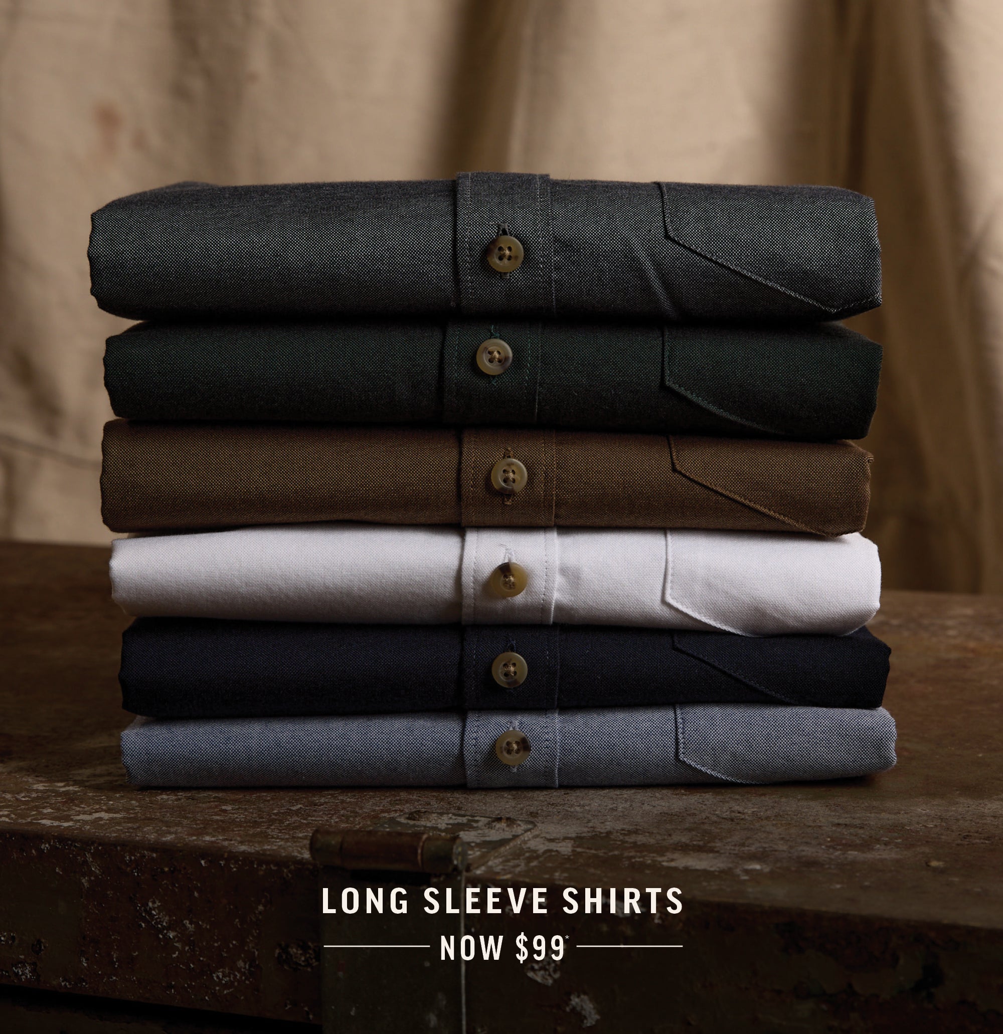 Six folded NZ long sleeve shirts in various colors stacked on a table, text reads Now $99.