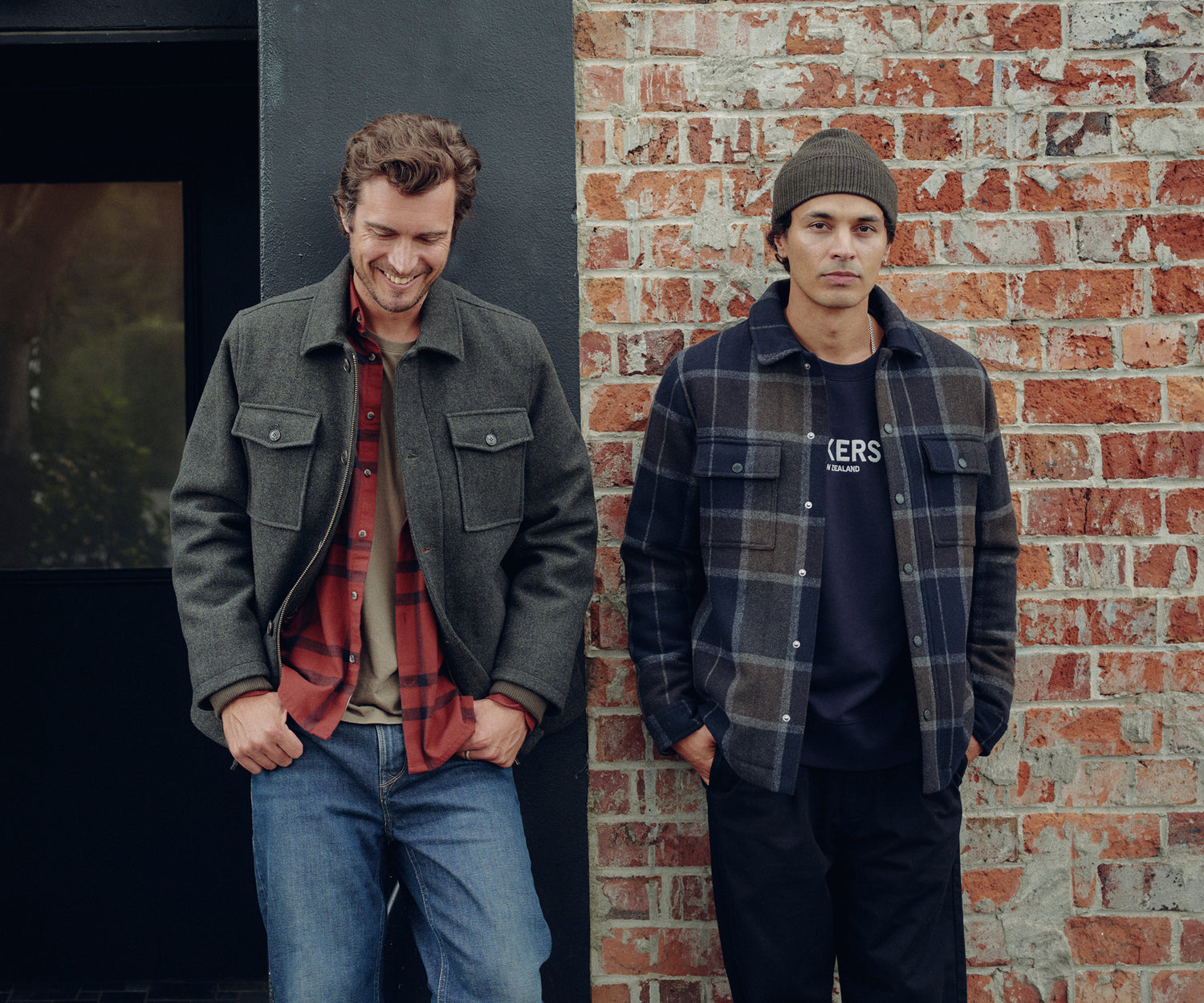 Two men stand in front of a brick wall. The man on the left smiles, wearing a gray jacket over a red plaid shirt and jeans. The man on the right looks serious, wearing a beanie, plaid jacket, and dark pants.