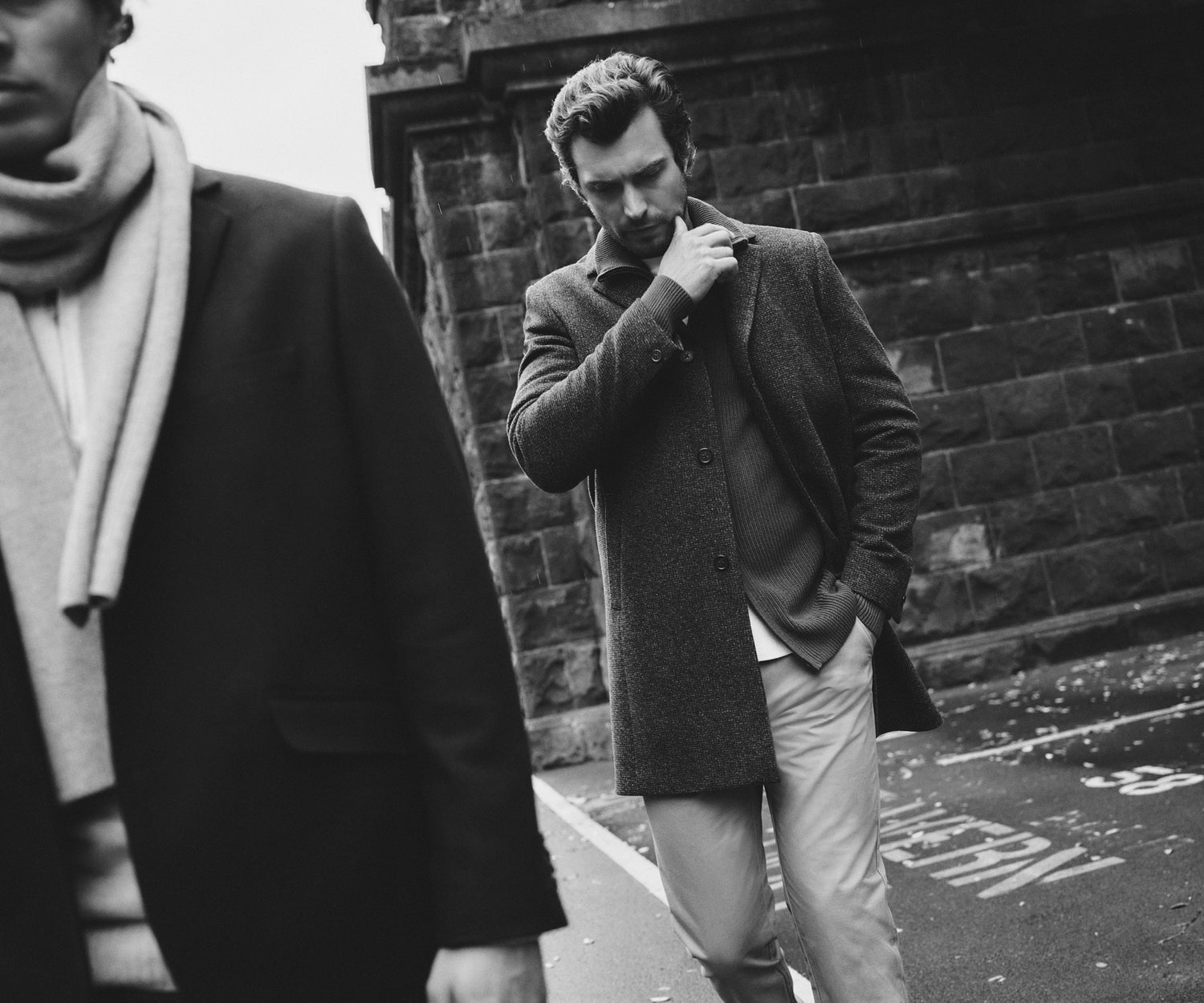 A black and white photo of two men walking outdoors. One man in a coat and scarf is partially visible in the foreground, while another man, wearing a coat and trousers, walks behind him near a stone wall, looking down pensively.